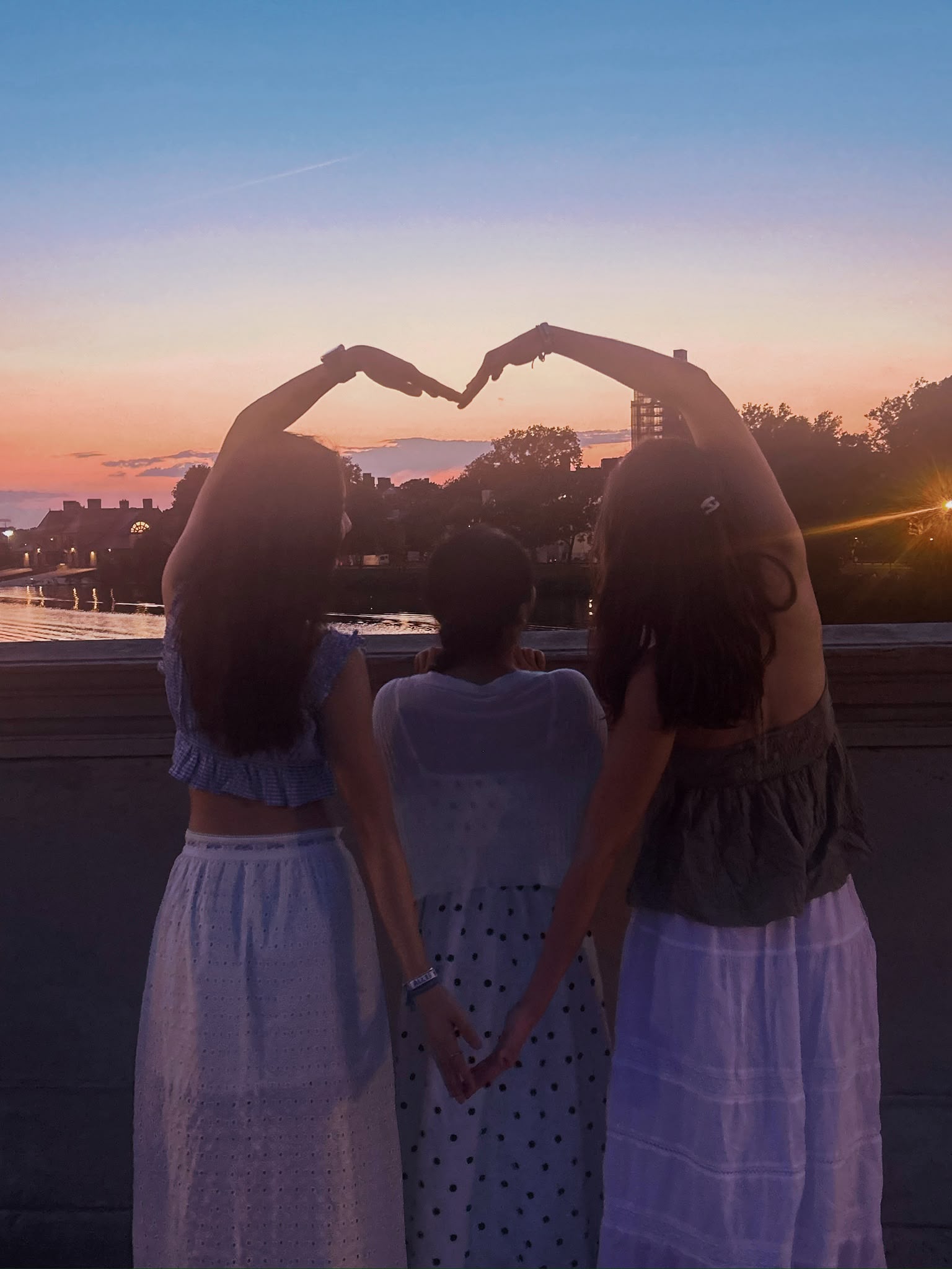 Three friends standing together at sunset, forming a heart shape with their arms